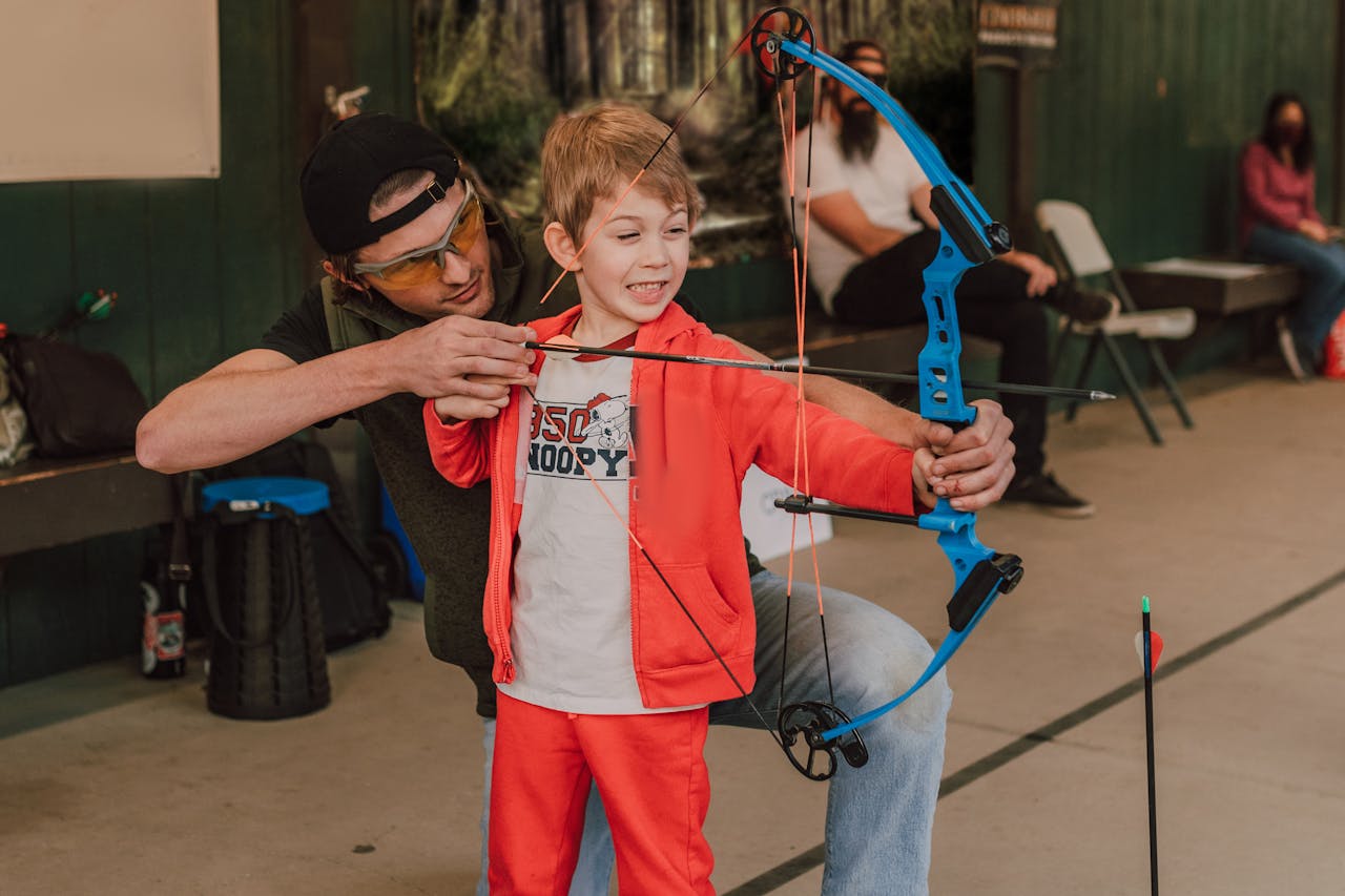 A young child learning archery with an instructor in an indoor setting, emphasizing fun and education.