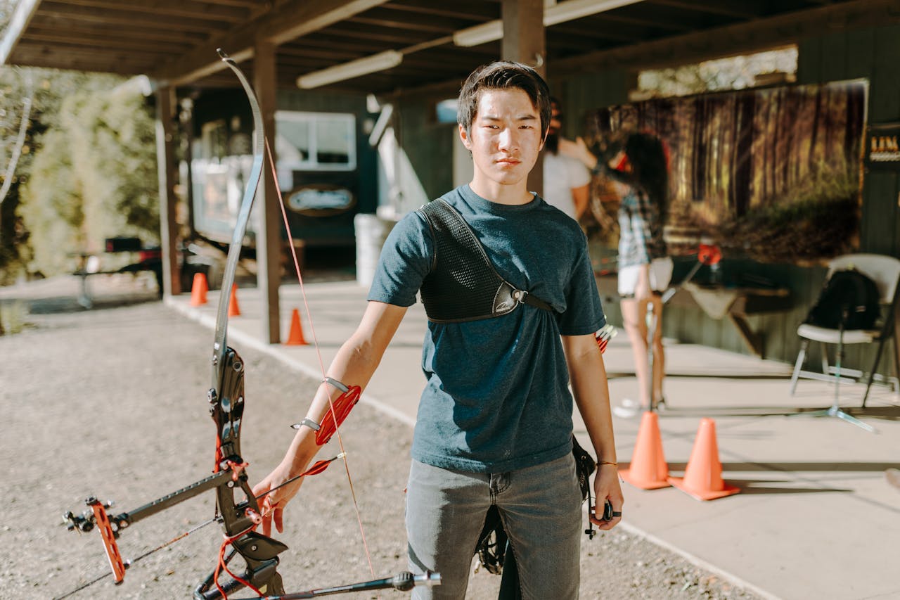 A young adult man practicing archery at an outdoor range on a sunny day.